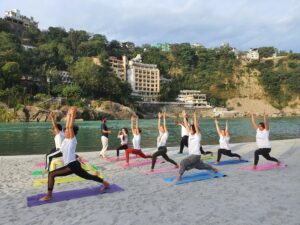 A group of adults practicing yoga on mats by a river with scenic hills in Rishikesh.